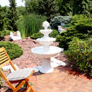 A white 3-tier outdoor fountain sits in a garden surrounded by trees and shrubs with water flowing down