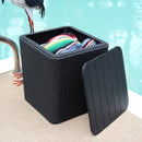 Black faux rattan storage bin with faux wood lid leaning against the side sitting pool side with a blanket colorful blanket inside.