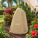 Beige faux rock utility cover in a tropical Florida garden beside a stucco home, surrounded by palm trees, bird-of-paradise plants, red hibiscus flowers, and lush green foliage.