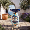 Colorful Mediterranean-style glass bird bath with a blue bowl and intricate blue, orange, and cream patterned pedestal, displayed on a stone patio surrounded by olive trees and lavender.