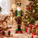 Decorative Santa figure in a green, burgundy, and gold outfit displayed indoors beside a decorated Christmas tree, wrapped gifts, and traditional holiday toys. The Santa stands on an ornate red and green base.