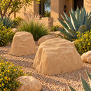 Two tan artificial landscape rocks in a Southwestern desert garden, surrounded by decorative pea gravel, agave plants, ornamental grasses, yellow flowering shrubs, and natural boulders outside a stucco home in warm sunlight.