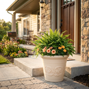 Cream-colored decorative planter filled with a lush mix of green foliage and peach flowers, displayed on a front porch beside a stone walkway.