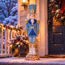 Blue, white, and gold Christmas nutcracker displayed outdoors in a snowy, decorated entryway, surrounded by evergreen garland, warm holiday lights, and a dark front door.