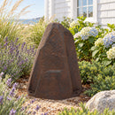 Dark brown faux rock utility cover with a tall, tapered shape and realistic stone texture, displayed in a landscaped garden with lavender, ornamental grasses, blooming hydrangeas, and a white house in the background.