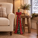 Green Christmas tree sculpture with a red bow and gold star displayed indoors beside an armchair, side table, candles, and holiday greenery.