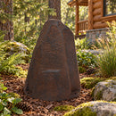 Dark brown faux rock utility cover with a tall, tapered shape and realistic stone texture, displayed in a woodland garden with ferns, moss-covered rocks, ornamental grasses, and a rustic wooden cabin in the background.