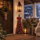 Green Christmas tree sculpture with a red bow and gold star displayed on a covered porch beside holiday greenery, lanterns, and a wreath.