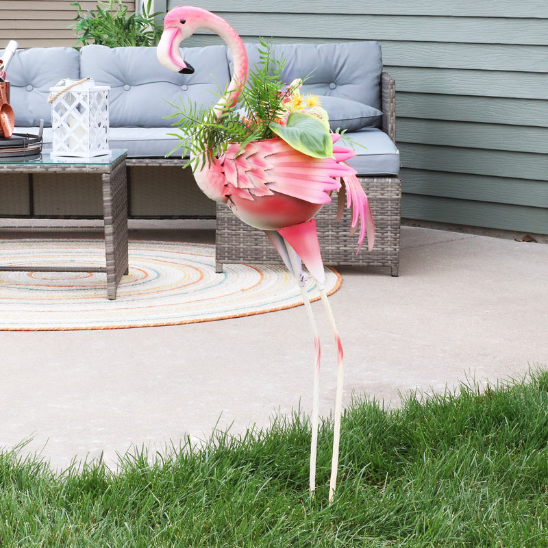 Outdoor flamingo garden statue with a planter full of flowers staked in the grass near an outdoor patio 