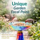 Vibrant multicolored glass birdbath filled with birdseed, with two birds perched on the bowl. Text highlights its unique garden focal point, vibrant colors, dramatic patterns, and handcrafted artisanal finish.