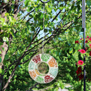 The colorful fly through bird feeder hangs from a metal stand outdoors with trees in the background.
