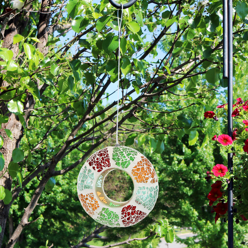 The colorful fly through bird feeder hangs from a metal stand outdoors with trees in the background.
