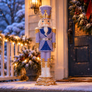 Purple, blue, white, and gold Christmas nutcracker standing outside a decorated home entrance, surrounded by snow-covered greenery and warm holiday lights.