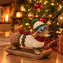 Winter dachshund statue wearing a knit hat, goggles, sweater, and scarf displayed beside a decorated Christmas tree with wrapped gifts.