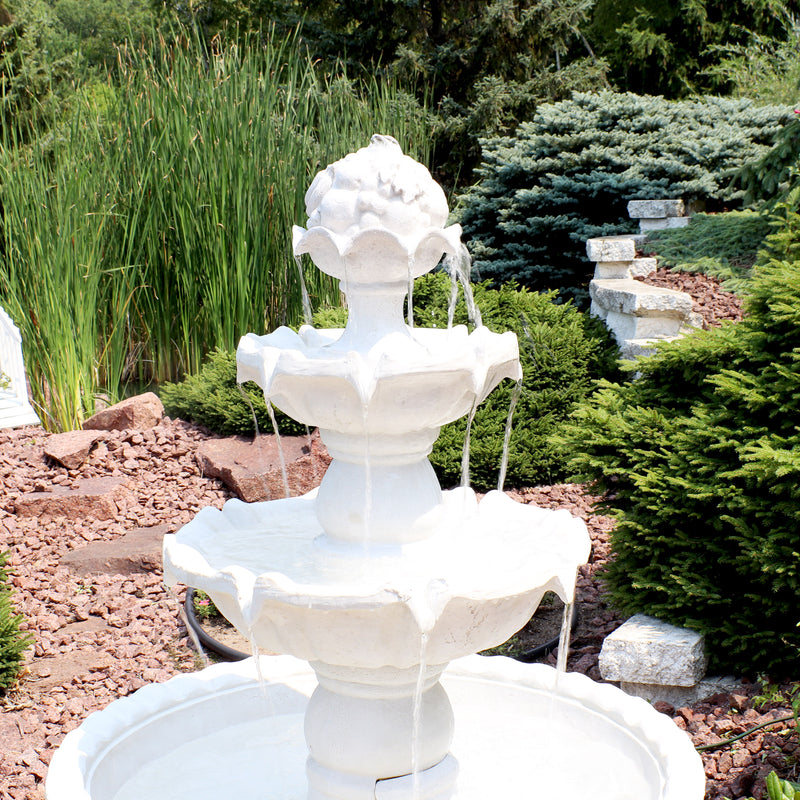 A white tiered outdoor fountain features a fruit design on top and has water pouring down the tiers in the yard.
