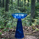 Blue glass birdbath displayed in a wooded garden setting, surrounded by trees and natural greenery.