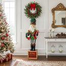 Green and red Christmas nutcracker statue holding a festive wreath and standing on a decorative “Merry Christmas” base in a holiday-decorated living room beside a Christmas tree, wrapped gifts, and a mantel with candles.