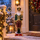 Decorative Santa figure in a green, burgundy, and gold outfit displayed outdoors on a snowy front porch beside illuminated holiday greenery and lanterns. The Santa stands on an ornate red and green base.