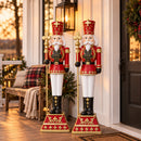 Two red and gold Christmas nutcracker kings holding gold scepters, displayed side by side on a covered front porch beside a black door, lantern, holiday wreath, and festive greenery.