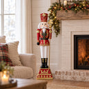 Red and gold Christmas nutcracker king holding a gold scepter, displayed indoors beside a decorated fireplace with illuminated garland, candles, and a cozy seating area.