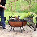 Copper fire pit with lit fire that is being sired by a fire poker while the spark screen rests on the left hand side of the fire pit.