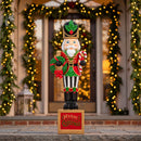 Green and red Christmas nutcracker statue holding a festive wreath and standing on a decorative “Merry Christmas” base in front of a warmly lit entryway decorated with illuminated holiday garland.