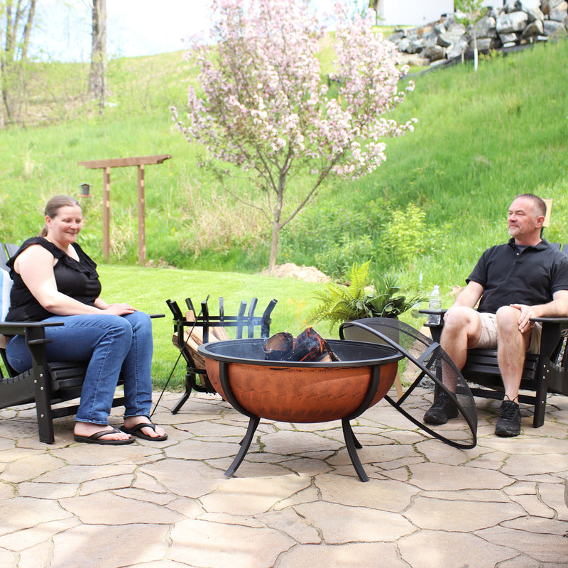 Woman and man sitting on black Adirondack chairs around a lit, copper-colored fire while the spark screen rests on the left-hand side.