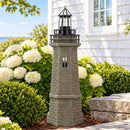 Weathered brown and green lighthouse statue displayed in a landscaped garden beside a coastal-style home, surrounded by white hydrangeas, green shrubs, and stone landscaping.