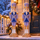 Two purple and blue Christmas nutcracker statues displayed side by side on a snow-covered front porch with illuminated holiday greenery and a decorated wreath.
