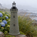Weathered brown and green lighthouse statue displayed on a rocky coastal landscape with blue hydrangeas, ornamental grasses, and an ocean view in the background.