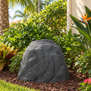 Dark gray faux rock cover with a realistic textured stone finish placed in a mulched garden bed surrounded by tropical plants, lush green shrubs, and bird of paradise flowers beside a stucco house.