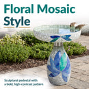 Mosaic Bloom Glass Bird Bath in indigo shown outdoors in a garden, featuring a sculptural pedestal with a bold blue, indigo, and aqua floral mosaic pattern and two small birds perched on the shallow bowl.