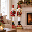Two red and gold Christmas nutcracker kings holding gold scepters, displayed side by side indoors beside a decorated fireplace with illuminated garland and a cozy seating area.