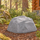 Gray decorative faux rock cover with a realistic stone texture and irregular shape, placed on a mulch-covered forest landscape surrounded by ferns, trees, and a rustic log cabin.