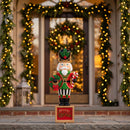 Festive Christmas nutcracker decoration dressed in a red, green, black, and gold uniform, holding a holiday wreath and candy cane while standing on a Merry Christmas base. Displayed on a stone walkway in front of a warmly lit entryway decorated with illuminated garlands and a Christmas wreath.