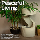 Cream-colored indoor tabletop fountain with a curved cascading design, shown among green plants and creating a gentle, calming water flow.