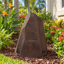 Dark brown faux rock utility cover with a tall, tapered shape and realistic stone texture, displayed in a tropical garden with palm trees, bird of paradise flowers, colorful foliage, and flowering plants beside a light-colored house.