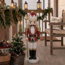 Red and green Christmas nutcracker king holding a gold scepter, displayed on a covered porch with holiday garland, potted greenery, lanterns, and a rustic wooden chair.