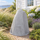 Light gray faux rock utility cover with a tall, tapered shape and realistic stone texture, displayed in a coastal garden with ornamental grasses, lavender, blooming hydrangeas, and a white house overlooking the water.