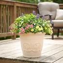 Cream-colored decorative planter filled with vibrant pink, yellow, and purple flowers and lush green foliage, displayed on a wooden deck beside outdoor seating.