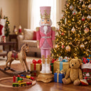 Pink, white, and gold Christmas nutcracker displayed indoors beside a decorated Christmas tree, wrapped gifts, a rocking horse, toys, and a teddy bear.