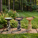 Three decorative pedestal bird baths in black, navy blue, and tan with cream patterns, displayed in a garden with birds perched on the rims and surrounded by flowers and greenery.