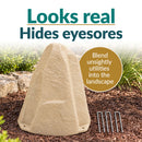 Beige faux rock utility cover with a realistic stone texture displayed in a landscaped garden. Text highlights its natural appearance and ability to conceal unsightly utilities. Four metal ground stakes are shown beside the rock.