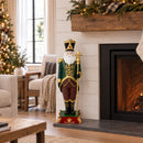 Decorative Santa figure in a green, burgundy, and gold outfit displayed indoors beside a lit fireplace and decorated Christmas tree. The Santa stands on an ornate red and green base.