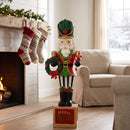 Green and red Christmas nutcracker statue holding a festive wreath and standing on a decorative holiday base in a cozy living room with a fireplace, stockings, and Christmas greenery.
