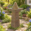 Outdoor pedestal fountain with a tiered pyramid water feature flowing into a square basin, surrounded by a colorful garden and stone pathway.