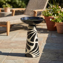 Black and cream decorative pedestal bird bath displayed on a stone patio, surrounded by outdoor furniture, greenery, and potted flowers.