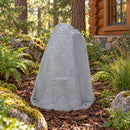 Light gray faux rock utility cover with a tall, tapered shape and realistic stone texture, displayed in a woodland garden with ferns, moss-covered rocks, ornamental grasses, and a rustic log cabin in the background.