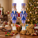 Two purple and blue Christmas nutcracker statues displayed side by side in a festive living room beside a decorated Christmas tree, wrapped gifts, a rocking horse, toy train, and teddy bear.