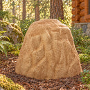Tan decorative faux rock cover with a tall, tapered shape and realistic stone texture, placed in a mulch-covered forest landscape surrounded by ferns, trees, and a rustic log cabin.